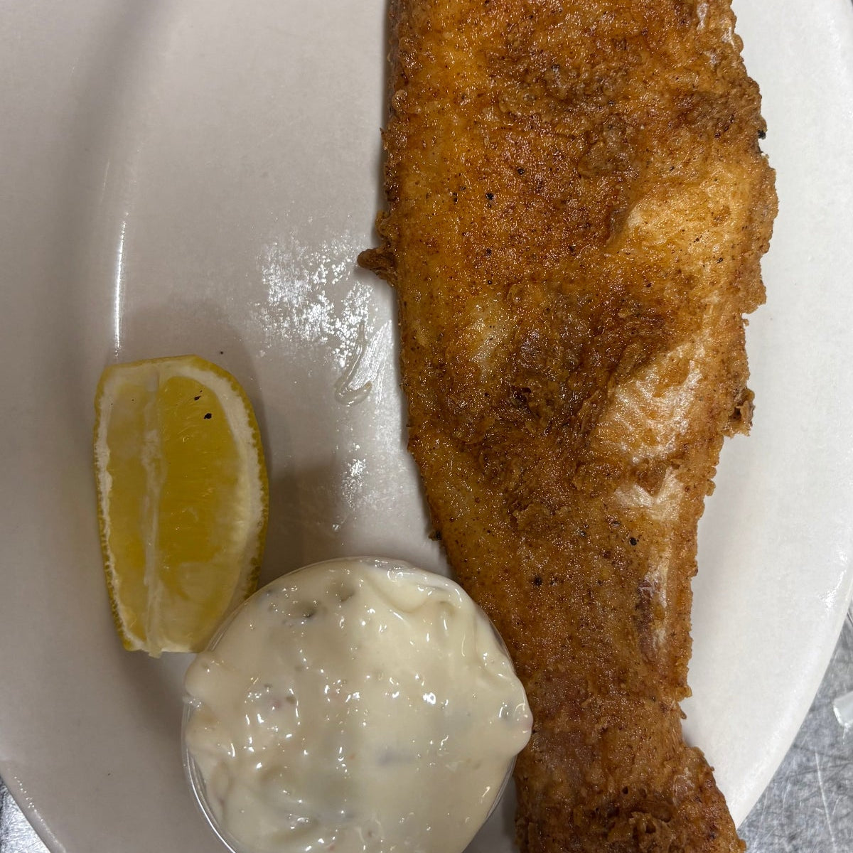 A whole fried fish served on a white oval plate with a lemon wedge and a dollop of white tartar sauce, photographed on a metallic surface.