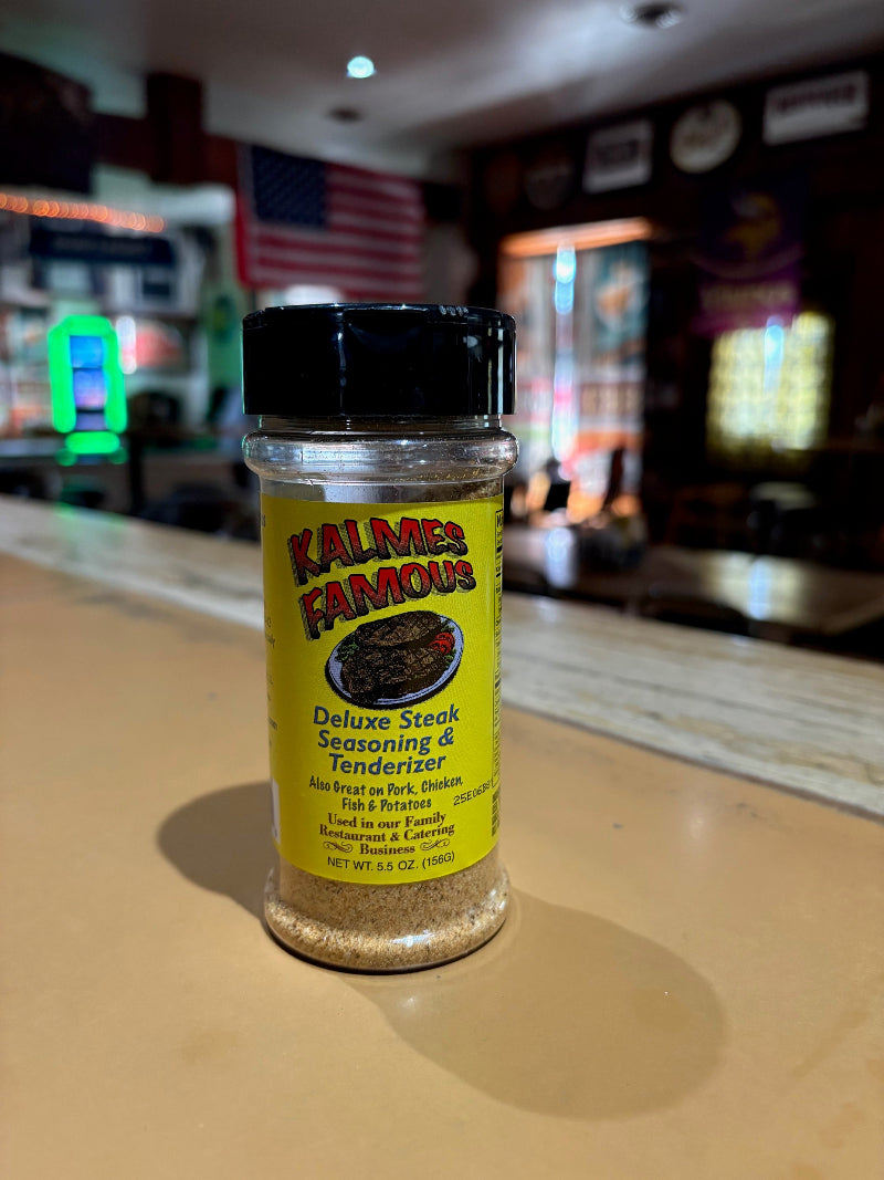 Bottle of Kalmes Famous 2-in-1 Steak Seasoning & Meat Tenderizer on a counter, with a restaurant interior softly blurred behind it.